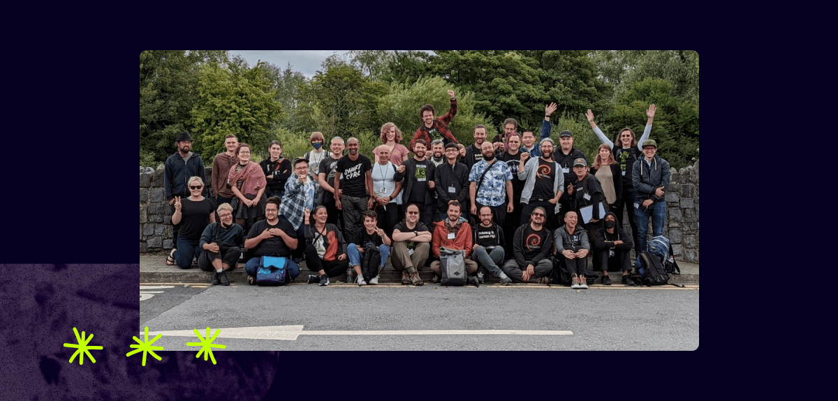 Staff and friends of Tor pose together in a large group outside on a cloudy day. People are making fun gestures, with some raising their hands towards the sky.